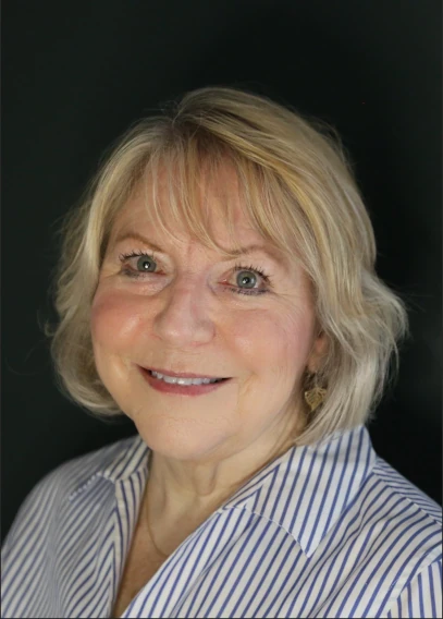 On a black background, Caucasian woman with short blonde hair, smiling with rosy cheeks. Wearing a white with blue striped blouse.